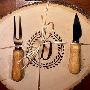 May include: A wooden cheese board with a burnt-in design of a wreath and the letter "D". A cheese knife and a cheese fork with wooden handles are tied to the board with a gold ribbon.