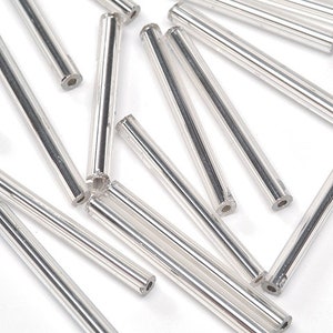May include: A collection of silver glass beads, each with a smooth, cylindrical shape. The beads are approximately 1 inch in length and have a reflective, metallic finish.