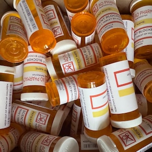 Può includere: Una collezione di flaconi di medicinali arancioni con etichette e tappi bianchi. Le etichette includono testo come "AO Pharmacy", "Nome del paziente" e "Prendere 1 compressa". I flaconi sono in una scatola di cartone.