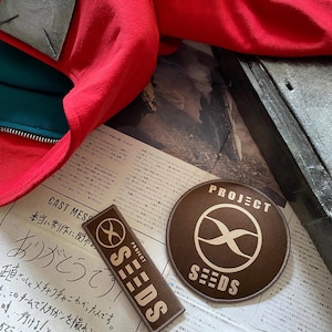 Op de afbeelding: Twee bruine stoffen patches met de tekst "PROJECT SEEDS" en een gestileerd oneindigheidssymbool. De patches liggen op een krant met Japanse tekst.