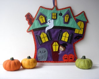 Könnte beinhalten: Ein Filz-Halloween-Haus mit einem violetten Körper, einem grünen Dach und orangefarbenen Verzierungen. Das Haus hat Fenster, eine Tür, einen Geist, eine Spinne und einen Grabstein mit der Aufschrift "R.I.P.". Vor dem Haus stehen drei kleine Kürbisse.