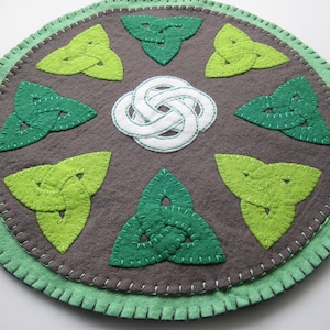 Peut inclure: Un dessous de plat rond en feutre vert et blanc avec un motif de nœud celtique. Le dessous de plat a une bordure extérieure verte et un cercle intérieur gris avec six nœuds celtiques verts et un nœud celtique blanc au centre.