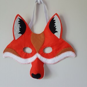 May include: A red felt fox mask with white felt accents around the eyes and ears. The mask has black felt eyes and a black felt nose.