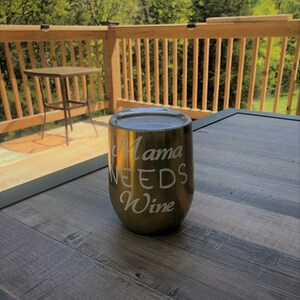 May include: A gold-colored stainless steel wine tumbler with the text "Mama Needs Wine" engraved on the side. The tumbler is sitting on a wooden table.