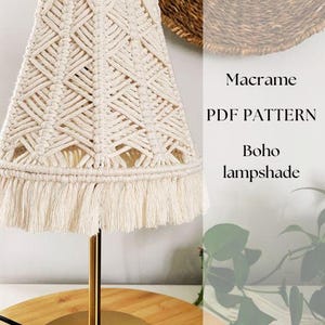 May include: A cream-colored macrame lampshade with a geometric pattern and fringe detail. The lampshade sits on a gold-colored metal base with a round wooden surface. Text on the image reads "Macrame PDF PATTERN Boho lampshade."