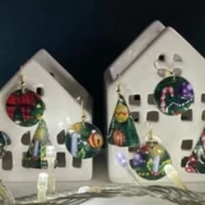Puede incluir: Casas blancas de cerámica con pendientes con temática de árbol de Navidad colgando de ellas. Los pendientes son redondos y presentan varios diseños navideños, incluyendo un bastón de caramelo, un árbol de Navidad y una estrella.
