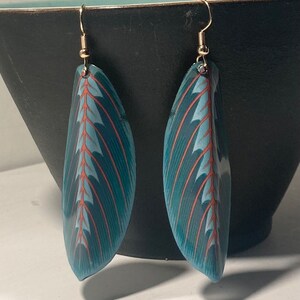 Puede incluir: Un par de pendientes de color azul turquesa con un patrón de hoja roja y blanca. Los pendientes están hechos de arcilla polimérica y tienen ganchos dorados.