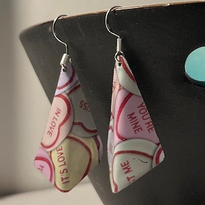May include: A pair of silver earrings with a triangular shape. The earrings are decorated with a pattern of pink and white hearts with various messages, including "You're Mine", "It's Love", and "In Love".