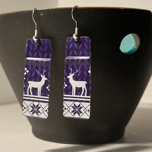 May include: Purple and white patterned earrings with a white reindeer silhouette. The earrings are rectangular and have a silver hook.