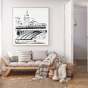 May include: A black and white line drawing of a city skyline with a bridge and a tall building in the center. The drawing is framed and hanging on a white wall above a light gray couch with throw pillows and a blanket.