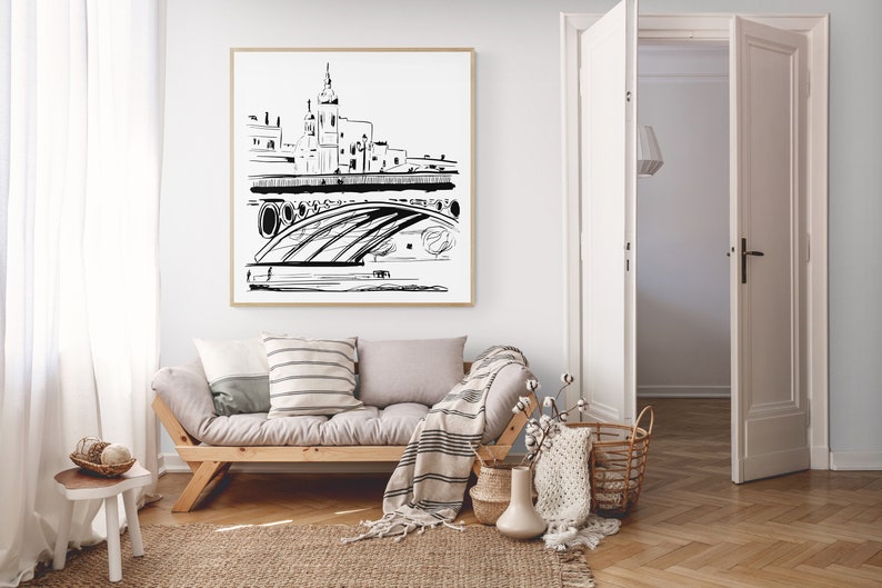 May include: A black and white line drawing of a city skyline with a bridge and a tall building in the center. The drawing is framed and hanging on a white wall above a light gray couch with throw pillows and a blanket.