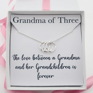 Puede incluir: Una caja de regalo blanca con un collar de plata en su interior. El collar tiene tres anillos entrelazados y una tarjeta que dice "Abuela de tres. El amor entre una abuela y sus nietos es para siempre."