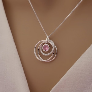 Pode incluir: Colar com pingente de prata com três círculos concêntricos. Uma pedra preciosa rosa está no centro, rodeada por um círculo de pequenas pedras transparentes. O colar pende de uma fina corrente de prata.