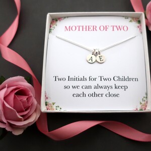 Peut inclure: Une boîte cadeau blanche contenant un collier en argent. Le collier est orné de deux breloques en argent avec les lettres A et E. La boîte est décorée d'un motif floral rose et porte l'inscription "MOTHER OF TWO" sur le devant. Le texte sous les breloques indique "Two Initials for Two Children so we can always keep each other close."