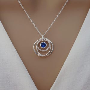 Op de afbeelding: Zilveren hangerketting met drie in elkaar grijpende cirkels. De middelste cirkel bevat een helderblauwe edelsteen, omringd door kleinere, heldere stenen. De ketting hangt aan een fijne zilveren ketting, wat een elegante uitstraling geeft.