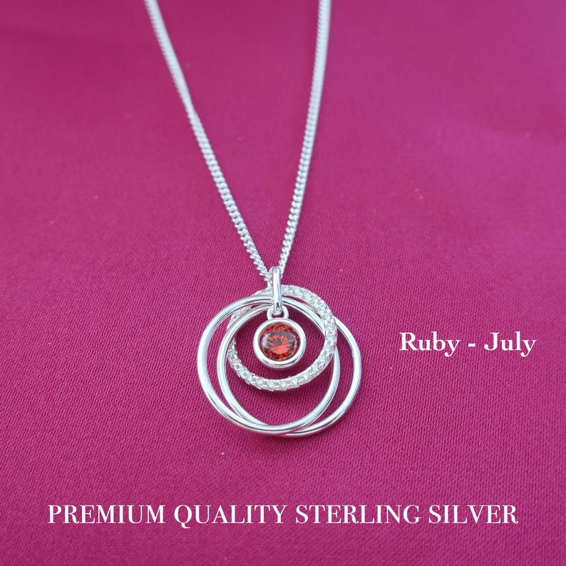 May include: A sterling silver necklace featuring a pendant with three interlocking circular rings, one of which is encrusted with small clear stones and holds a central round red ruby stone. The text reads Ruby - July and PREMIUM QUALITY STERLING SILVER.