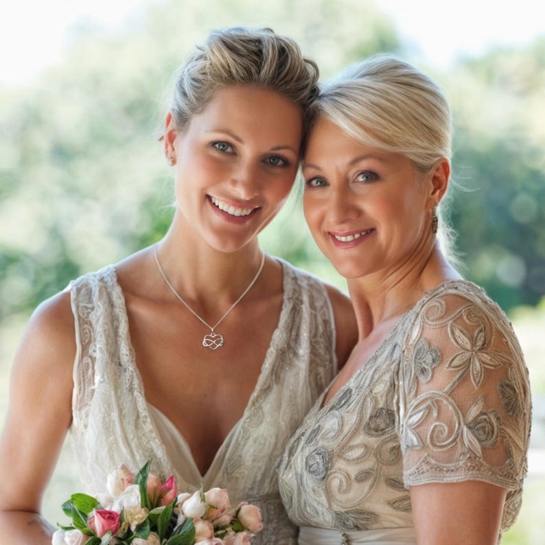May include: Two women, a bride and her mother, smiling at the camera. The bride is wearing a white lace dress and a silver necklace with a heart pendant. Her mother is wearing a silver sequined dress with a lace overlay. They are both standing in front of a blurred green background.