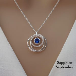 May include: A silver necklace featuring a pendant with three interlocking circular rings. The center ring is set with a round, deep blue sapphire gemstone. The text Sapphire September is printed in black in the lower right corner of the image.