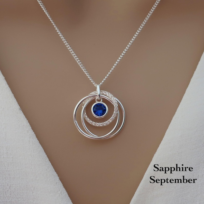 May include: A silver necklace featuring a pendant with three interlocking circular rings. The center ring is set with a round, deep blue sapphire gemstone. The text Sapphire September is printed in black in the lower right corner of the image.