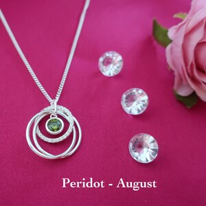May include: A silver necklace featuring a pendant with three interlocking rings, one of which is encrusted with small clear stones and another holding a single round green peridot stone. The text Peridot - August is printed below.