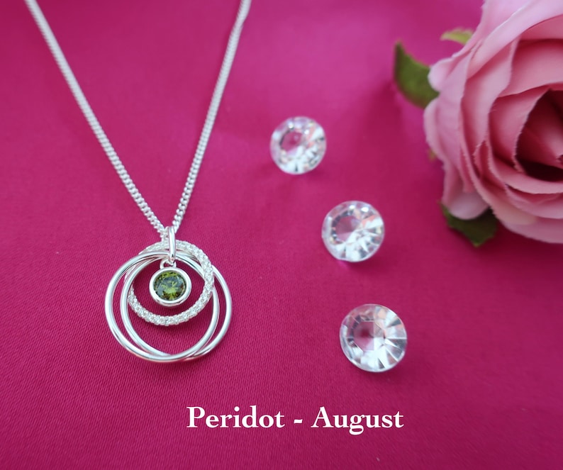 May include: A silver necklace featuring a pendant with three interlocking rings, one of which is encrusted with small clear stones and another holding a single round green peridot stone. The text Peridot - August is printed below.