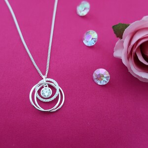 May include: A silver-toned necklace featuring a pendant with three interlocking circular rings, one of which is encrusted with small clear stones, and a central round clear stone setting. The necklace is displayed alongside three loose iridescent stones.