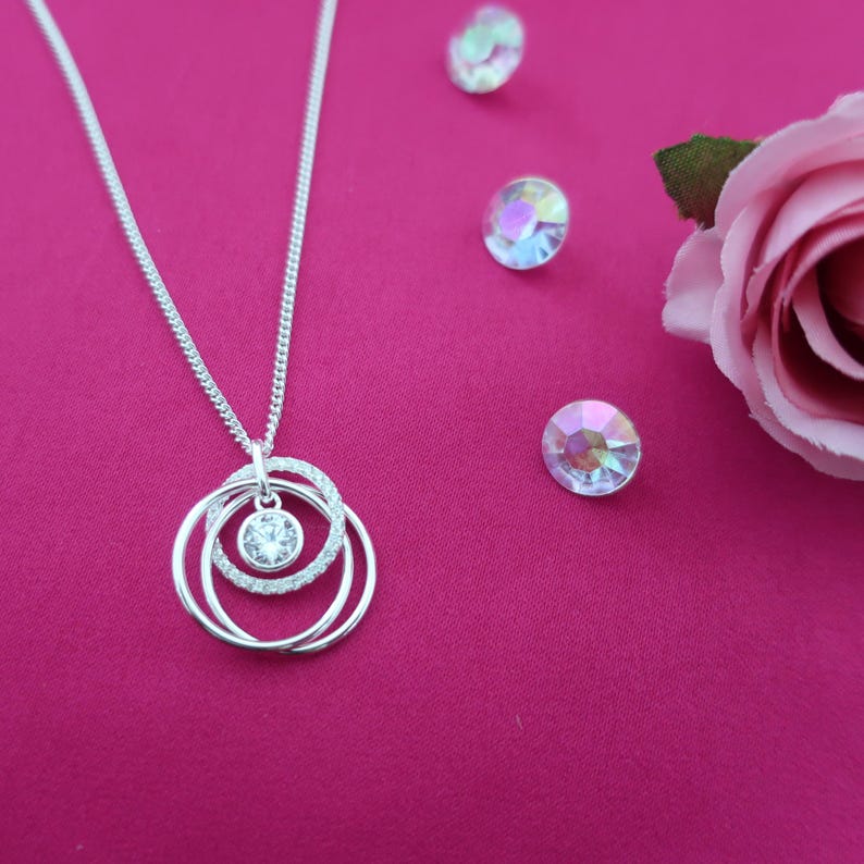 May include: A silver-toned necklace featuring a pendant with three interlocking circular rings, one of which is encrusted with small clear stones, and a central round clear stone setting. The necklace is displayed alongside three loose iridescent stones.