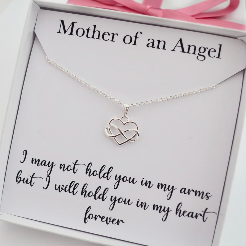 May include: A white box with a silver necklace featuring an infinity heart charm. The box has the text "Mother of an Angel" and the quote "I may not hold you in my arms but I will hold you in my heart forever".