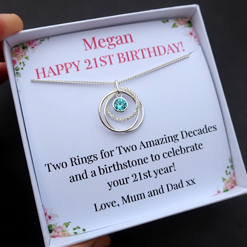 May include: A silver necklace with a pendant featuring two interlocking rings, one plain and one encrusted with small clear stones, centered with a single round bright blue gemstone. The necklace is presented in a white gift box with text.
