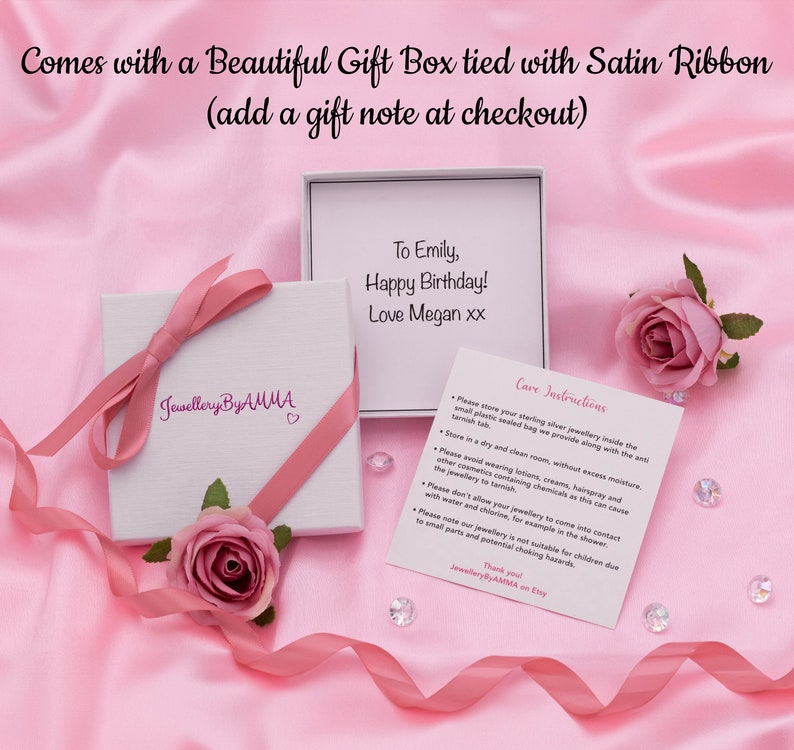May include: A white gift box with a pink satin ribbon tied around it. The box has a pink heart and the words "Jewellery By AMMA" printed on it. A card with the text "To Emily, Happy Birthday! Love Megan xx" is visible in the background. The card is on a pink surface.