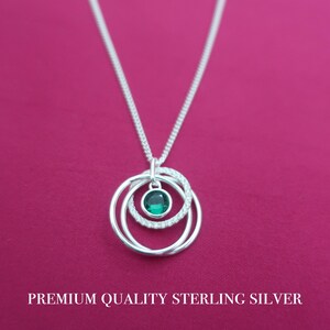 May include: A sterling silver necklace featuring a pendant with three interlocking circular rings. One ring is set with a round, faceted green gemstone, while another is encrusted with small clear stones. The text reads PREMIUM QUALITY STERLING SILVER.