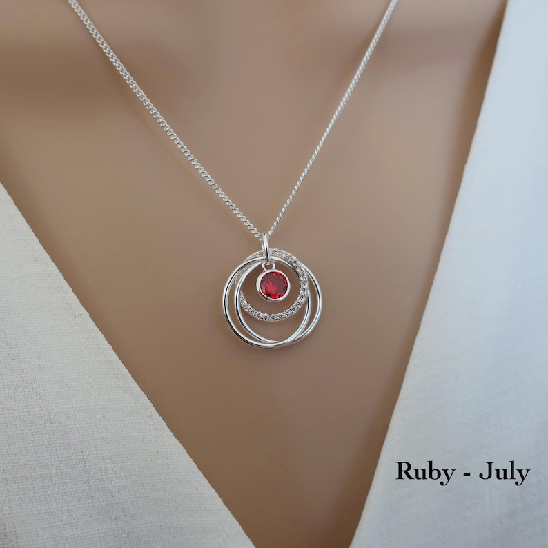 May include: A silver necklace featuring a pendant with three interlocking circular rings, one of which is encrusted with small clear stones and another holding a single round red gemstone. The text Ruby - July is printed in the bottom right corner.