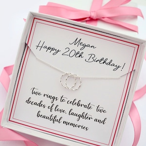 May include: A white gift box with a pink ribbon and a silver necklace with two intertwined rings. The box has a card inside that reads "Happy 20th Birthday! Megan" and "Two rings to celebrate two decades of love, laughter and beautiful memories".