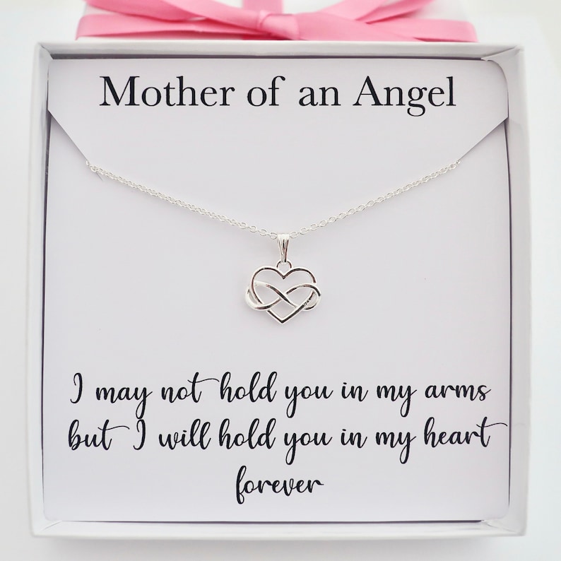 May include: A silver necklace with an infinity symbol inside a heart pendant. The necklace comes in a white box with a pink ribbon and a card that reads "Mother of an Angel" and "I may not hold you in my arms but I will hold you in my heart forever".