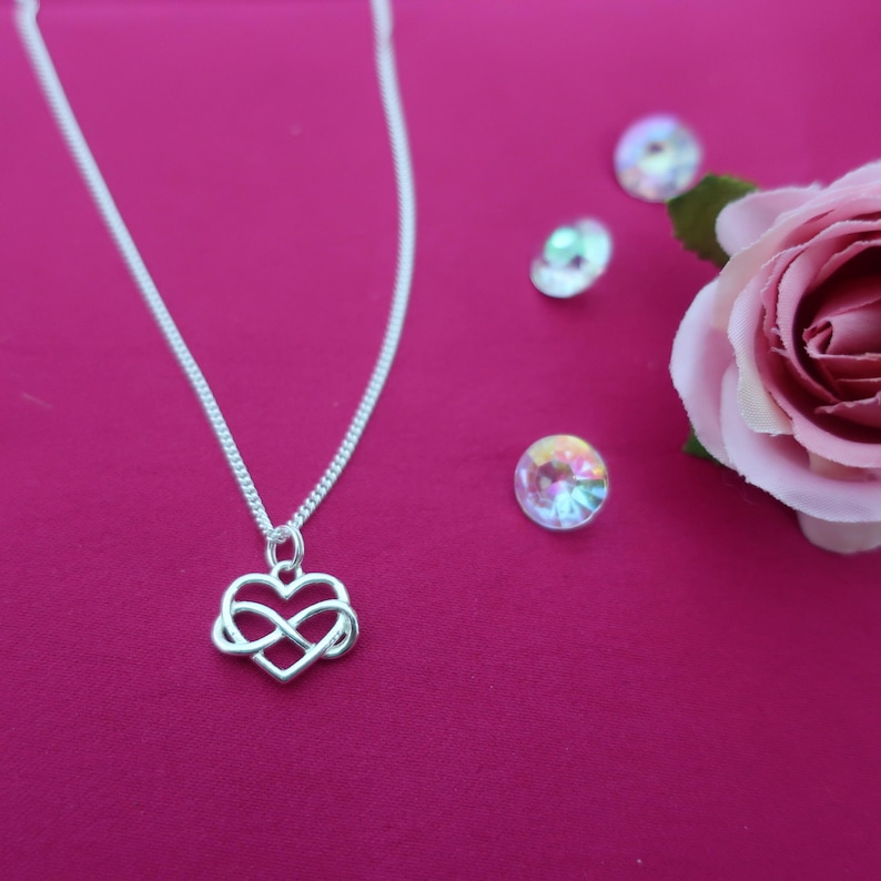 May include: A silver necklace with an infinity symbol heart pendant. The necklace is on a pink background with iridescent rhinestones and a pink rose.