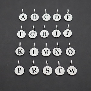 Puede incluir: Colección de colgantes redondos plateados con letras del alfabeto de la A a la W. Cada colgante tiene una letra diferente grabada. Están dispuestos en filas y columnas sobre un fondo oscuro. Cada colgante tiene un pequeño lazo en la parte superior.