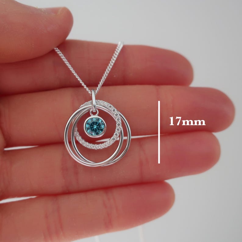 May include: A silver-toned necklace featuring a pendant with three interlocking circular rings. One ring is encrusted with small clear stones, and a central circular charm holds a single bright blue stone. The pendant measures 17mm in diameter.