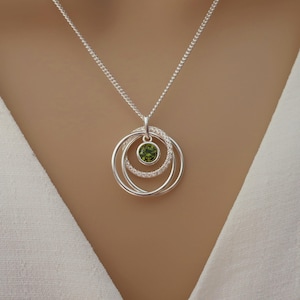 May include: A silver necklace featuring a pendant with three interlocking circular rings. The center ring is set with a round, faceted olive green gemstone, while one of the surrounding rings is encrusted with small, clear, sparkling stones.