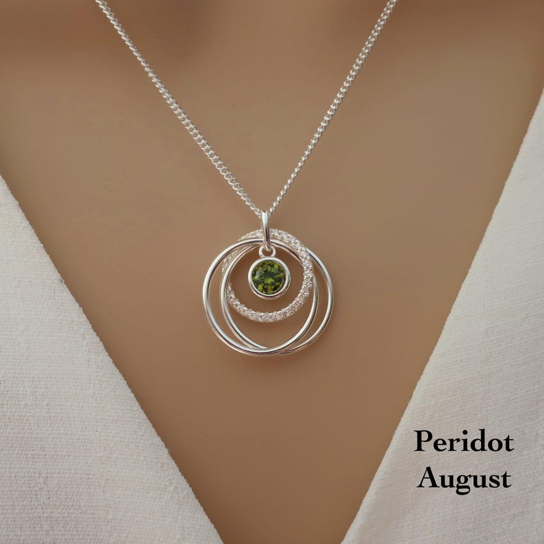 May include: A silver necklace featuring a pendant with three interlocking circular rings. The center ring is set with a round, faceted green peridot gemstone. The text Peridot August is printed in the lower right corner of the image.