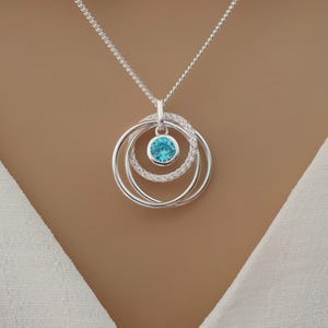 May include: A silver-toned necklace featuring a pendant with three interlocking circular rings. One ring is encrusted with small clear stones, while another holds a single, round, bright blue gemstone in a bezel setting at the center.