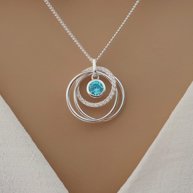 May include: A silver-toned necklace featuring a pendant with three interlocking circular rings. One ring is encrusted with small clear stones, while another holds a single, round, bright blue gemstone in a bezel setting at the center.
