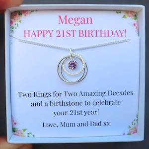 Op de afbeelding: Een zilveren ketting met een hanger met twee in elkaar grijpende ringen en een centrale paarse edelsteen, gepresenteerd in een geschenkdoos. De doos heeft de tekst "Megan HAPPY 21ST BIRTHDAY!" en "Two Rings for Two Amazing Decades".