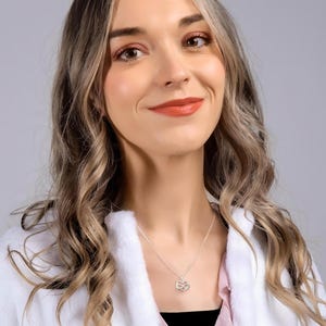 May include: A woman with long, wavy brown hair is wearing a white jacket and a pink shirt. She is smiling and looking to the right. She is wearing a silver necklace with a small infinity symbol pendant.