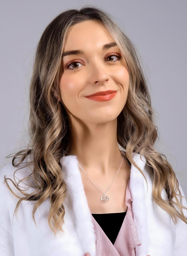May include: A woman with long, wavy brown hair is wearing a white jacket and a pink shirt. She is smiling and looking to the right. She is wearing a silver necklace with a small infinity symbol pendant.