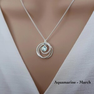 May include: A silver necklace featuring a pendant with three interlocking circular rings. The center ring is set with a single light blue round gemstone, while another ring is embellished with small clear stones. The text reads Aquamarine - March.