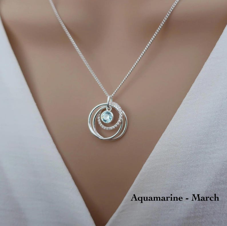 May include: A silver necklace featuring a pendant with three interlocking circular rings. The center ring is set with a single light blue round gemstone, while another ring is embellished with small clear stones. The text reads Aquamarine - March.
