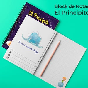 Puede incluir: Un cuaderno de espiral titulado El Principito con una portada de color morado oscuro con temática espacial. Las páginas abiertas muestran una ilustración de un elefante azul, el texto Lo esencial es invisible a los ojos y un personaje.