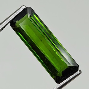 May include: A vibrant green gemstone with a rectangular cut and a faceted finish. The gemstone is held in a metal clamp.