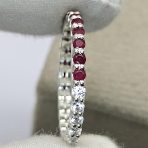 Könnte beinhalten: Ein schmaler Eternity-Ring mit einer Reihe abwechselnd rund geschliffener, tiefroter Rubine und klarer weißer Steine in einer silberfarbenen Metallfassung. Der Ring wird vertikal gehalten und zeigt die durchgehende kreisförmige Anordnung.