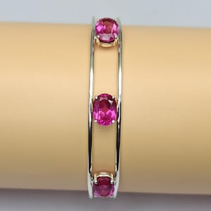 Peut inclure: Un bracelet manchette argenté composé de deux bandes métalliques parallèles reliées par trois pierres précieuses roses vives, de forme ovale et serties. Les pierres sont disposées verticalement au centre du bracelet ouvert.